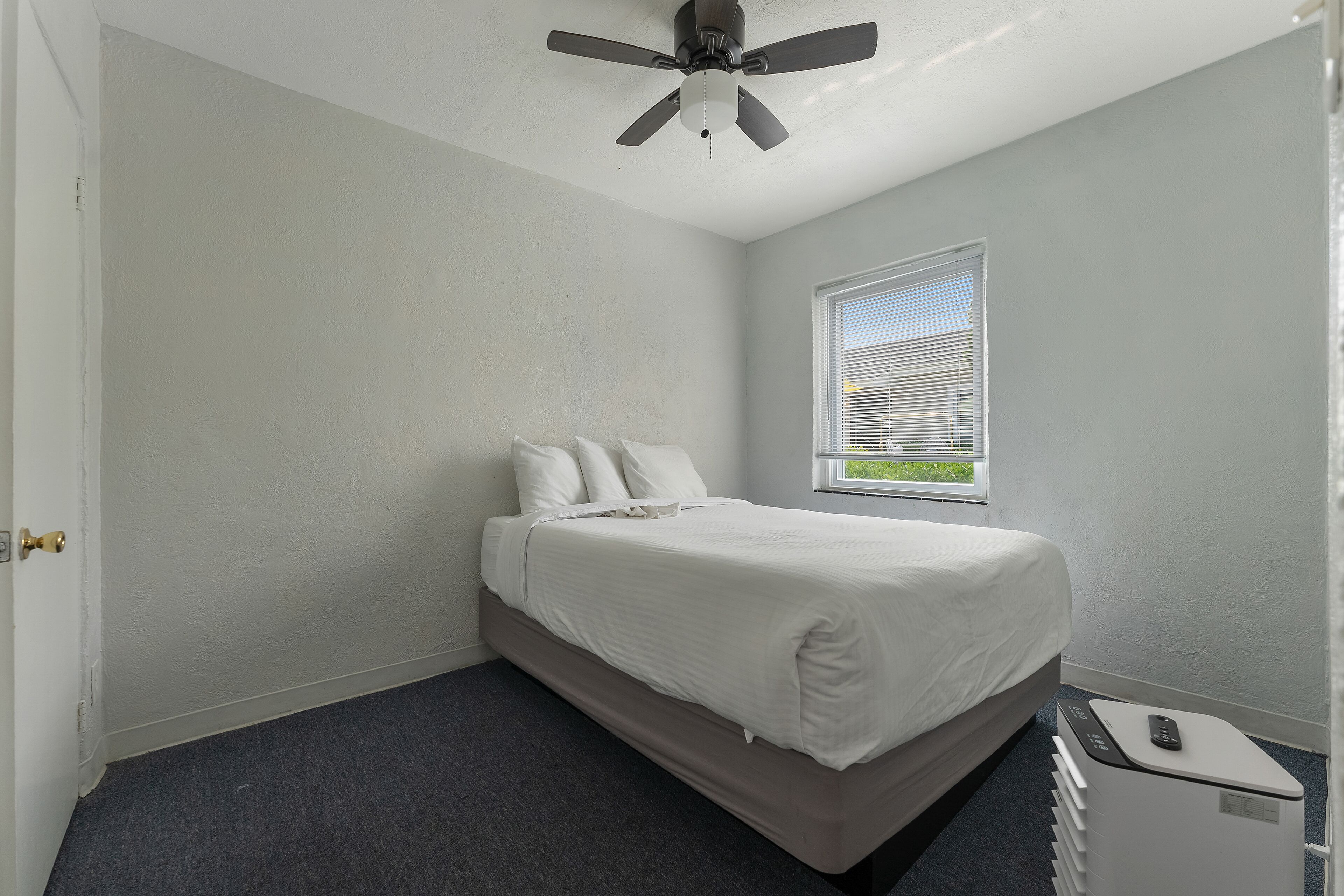 Un dormitorio con una cama, ventilador de techo, ventana y mesita de noche blanca.
