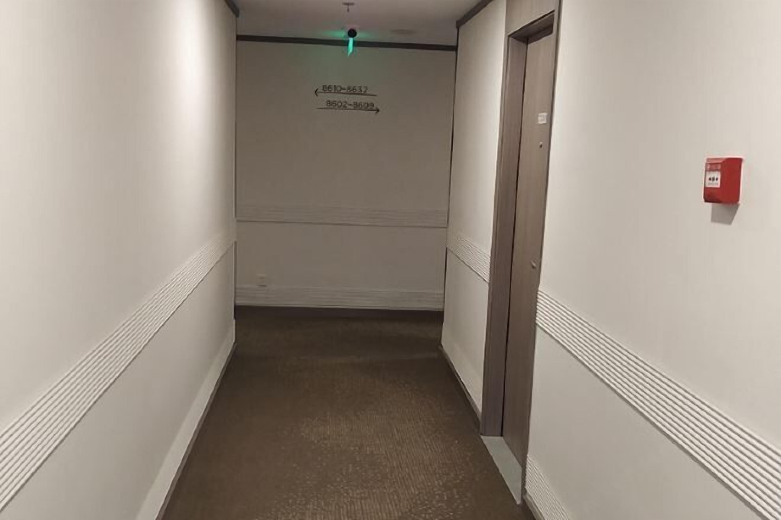 A narrow hallway with a carpeted floor, white walls, and a door on the right. There is a fire alarm on the wall and a green exit sign above the door.