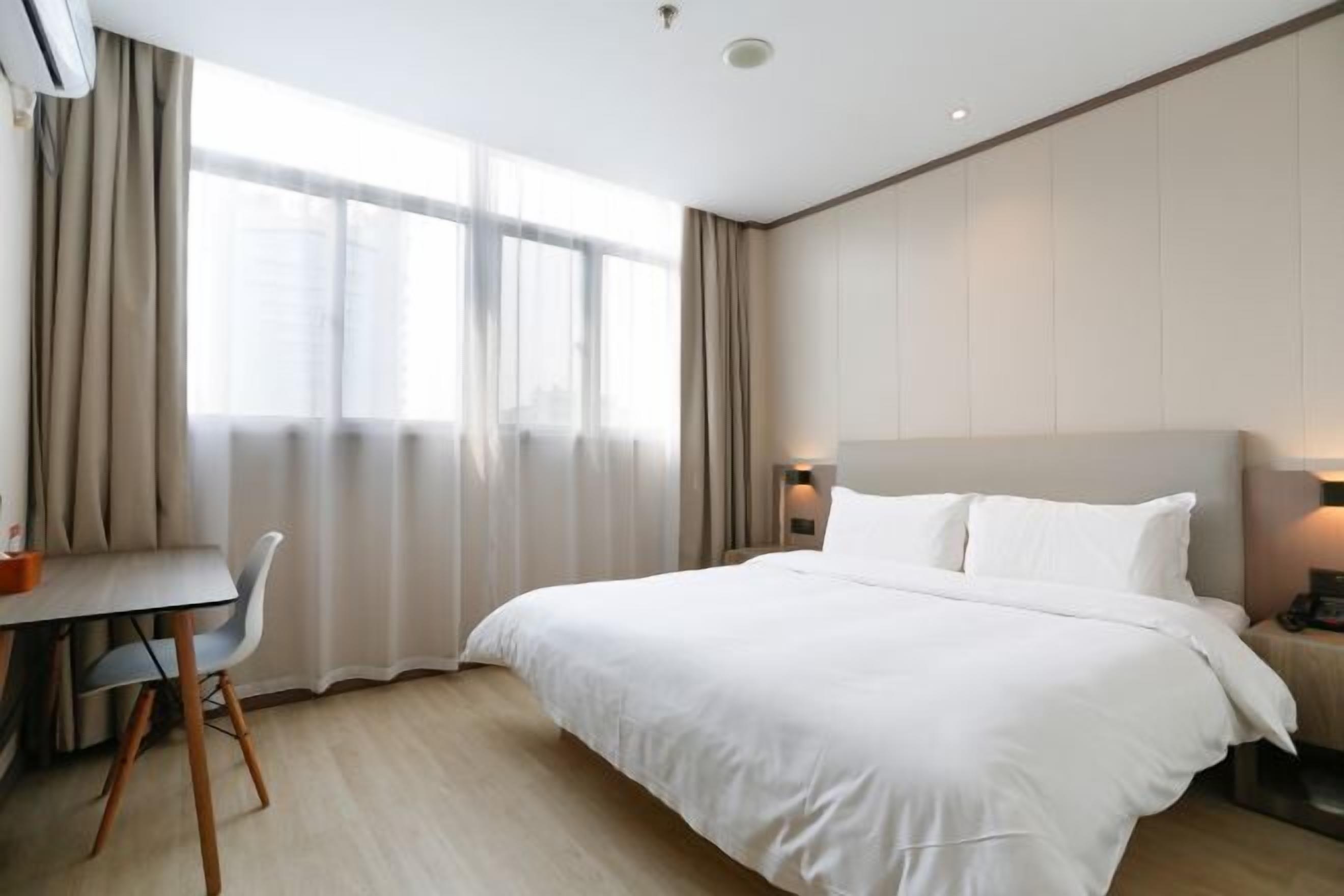 A hotel room with a large bed, a desk, and a chair. There are curtains on the windows, and a wall-mounted lamp is visible.
