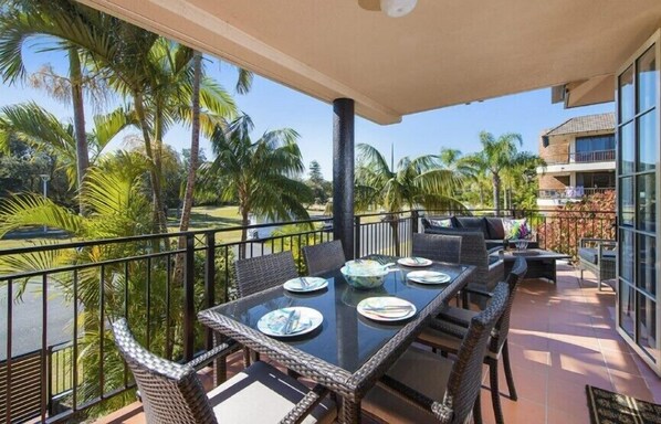 A balcony with a glass table set for dining, surrounded by wicker chairs, overlooking a palm tree-lined view. - Seabreeze 4 on Lawson Street (Byron Shire)