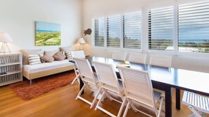 A spacious room with a large dining table, white chairs, a sofa, and a view of the outdoors through large windows with blinds. - 3 bedroom 2 bathroom spacious unit with water views. (Point Lookout)
