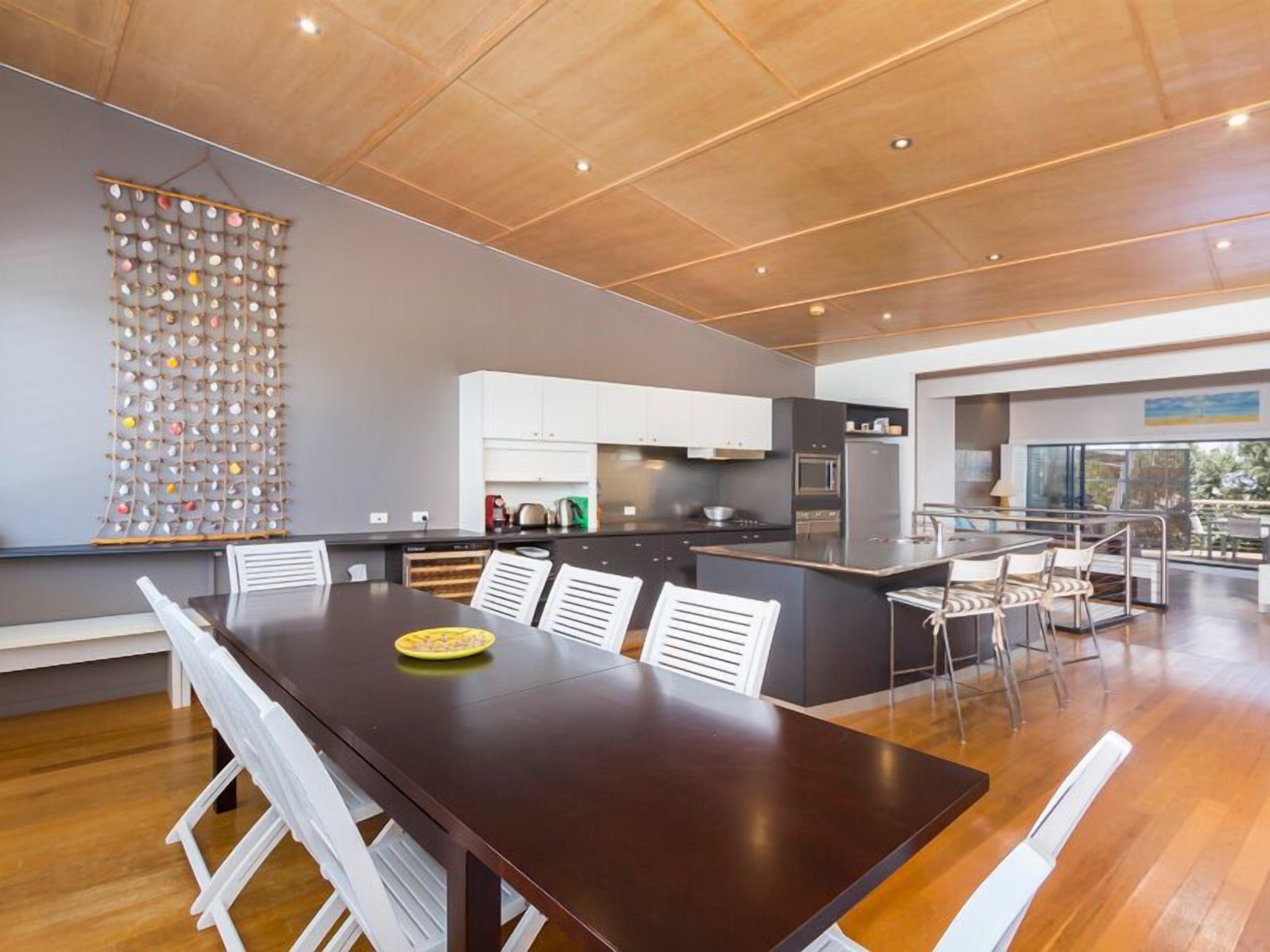 Una cocina moderna con armarios blancos, una isla central y un comedor con una mesa de madera oscura y sillas blancas.