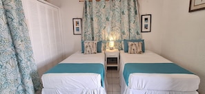 A room with two beds, a nightstand with a lamp, a window with curtains, and a door with a shutter. - Fully A/C Ground-Floor Oasis Steps from the Pool & South Coast (Bridgetown)