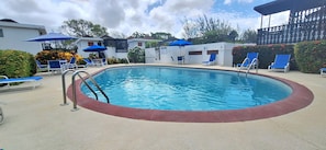 A swimming pool with blue lounge chairs and umbrellas, surrounded by white buildings and greenery.