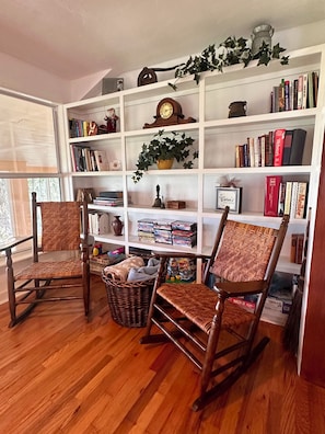 A room with a wooden floor, two rocking chairs, a bookshelf filled with books and decorative items, and a window with blinds. - Lake living close to Gainesville, UF Health and University of Florida (Melrose)