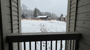 A view from a window with a wooden railing, looking out at a snowy landscape with a covered area and buildings in the distance. - Ski In - Ski Out Condo in OberTal 'F' Building! (Stratton Mountain)