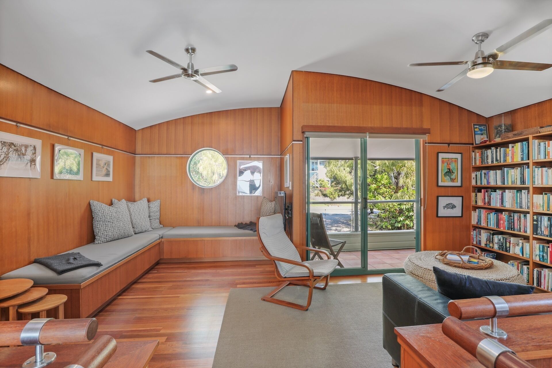 A modern living room with wooden paneling, a built-in bench, a rocking chair, a bookshelf, and two ceiling fans.