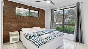 A bedroom with a brick wall, a bed with patterned bedding, a ceiling fan, and a large window with curtains. - Pets Allowed in this comfortable three bedroom house centrally located. (Point Lookout)