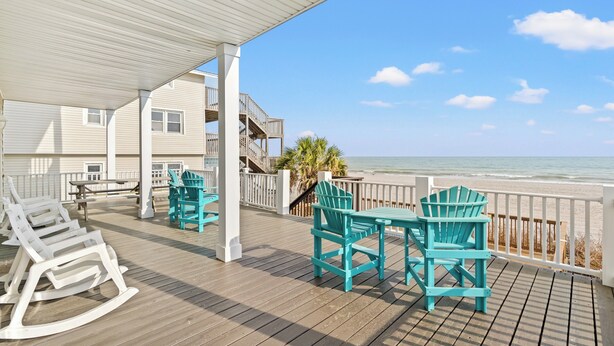 Una terraza de madera con mecedoras y una mesa, con vista a la playa.