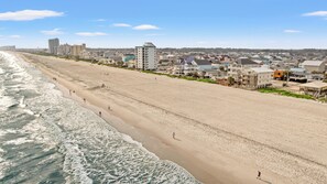 A wide sandy beach with ocean waves, beachfront buildings, and a clear sky.