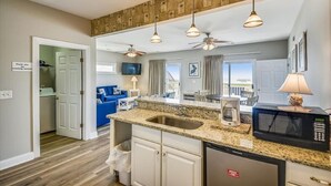 Cocina moderna con encimeras de granito, lavavajillas integrado y microondas. Hay un comedor con mesa y sillas, y vistas a la playa a través de la puerta abierta.