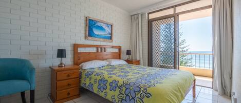A bedroom with a bed, two nightstands, a blue chair, a white brick wall, a painting, and a balcony with a view.