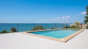 Una piscina rectangular con solárium empedrado, rodeada por una valla metálica, y vistas a un océano azul y despejado.