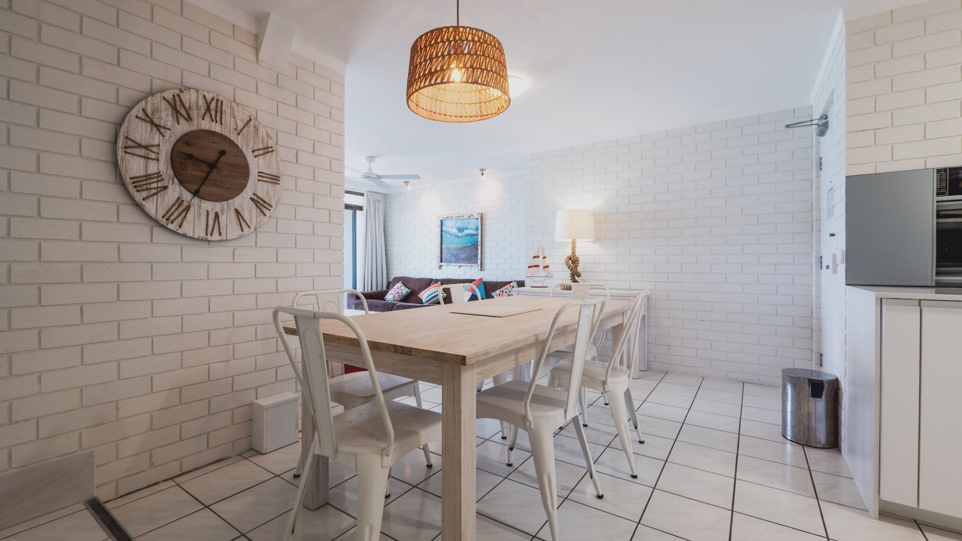 A modern kitchen with white brick walls, a wooden dining table with white chairs, a hanging light fixture, and a large wall clock.