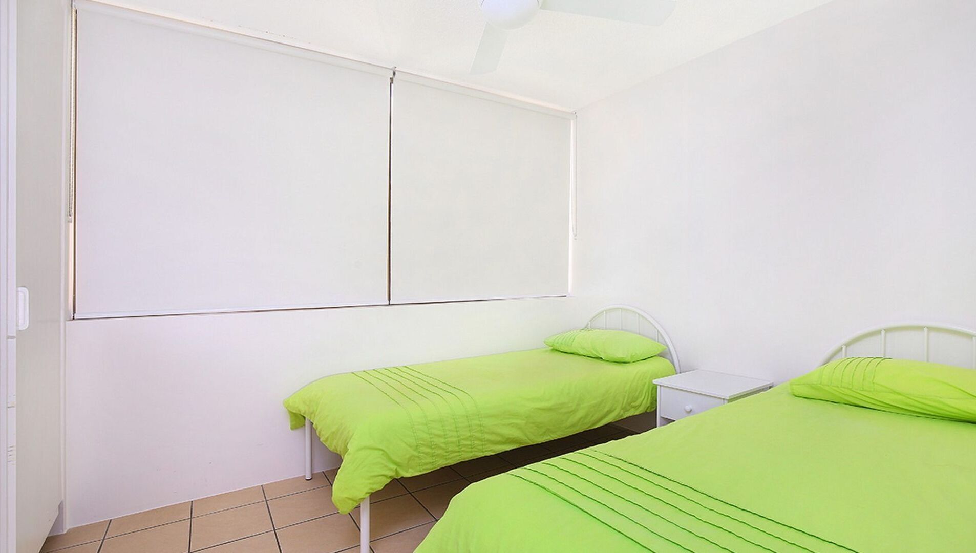 A room with two beds, each with green bedding, a white headboard, and a small bedside table.