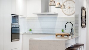 Ett modernt kök med vita skåp, en stor köksö med barstolar, en inbyggd ugn och hängande lampor.