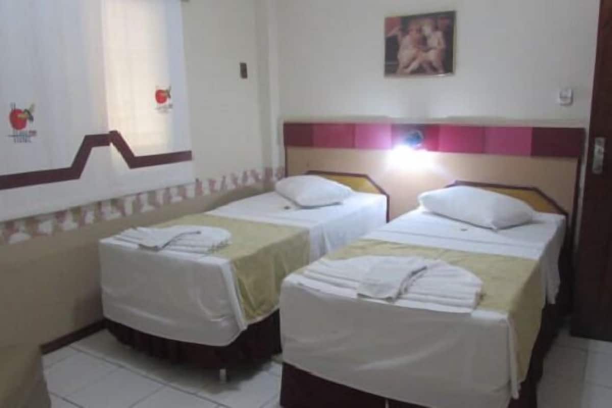 A hotel room with two single beds, each with a pillow and a folded towel. There is a wall-mounted picture and a window with curtains.