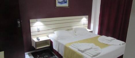 A hotel room with a bed, a bedside table, a nightstand, a telephone, a painting on the wall, and a red curtain.