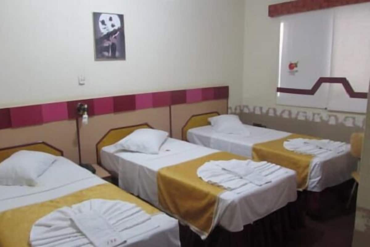 A hotel room with three single beds, each with a white bedspread and a yellow blanket. There is a wall-mounted light fixture and a framed picture above the beds.