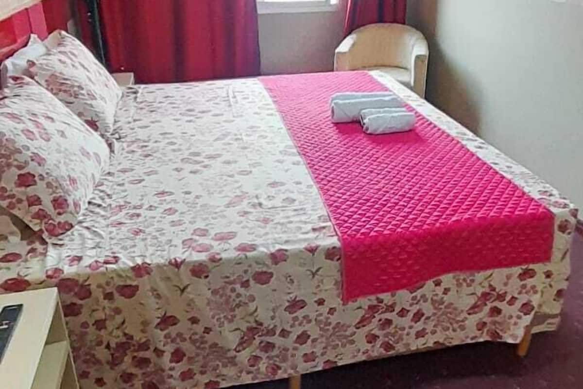 A single bed with a floral bedspread and a red blanket, a chair, a window with red curtains, and a wall-mounted TV.
