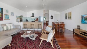 Amplia sala de estar con sofá, mesa de centro y mesa de comedor con sillas. Hay una zona de cocina con estufa, fregadero y refrigerador. La habitación tiene un techo alto con ventilador de techo.