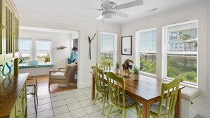 A bright dining area with a wooden table and chairs, a ceiling fan, and large windows with blinds.