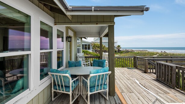 A covered outdoor deck with a table and chairs, overlooking a beach and ocean.