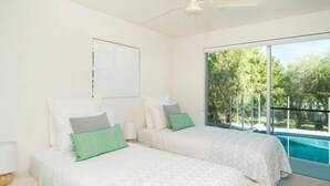 A bedroom with two beds, a ceiling fan, and a large window with a view of trees and a pool.