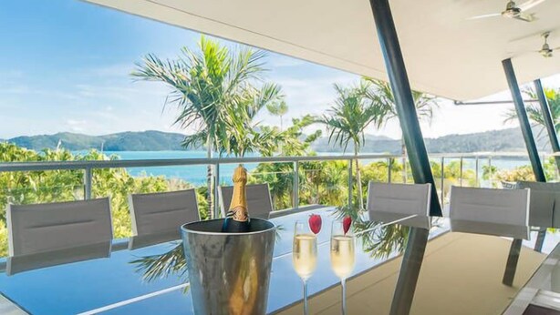 A table with a bottle of champagne, two glasses of wine, a plate with grapes and cheese, and a bucket, set against a scenic view of a lake and mountains.