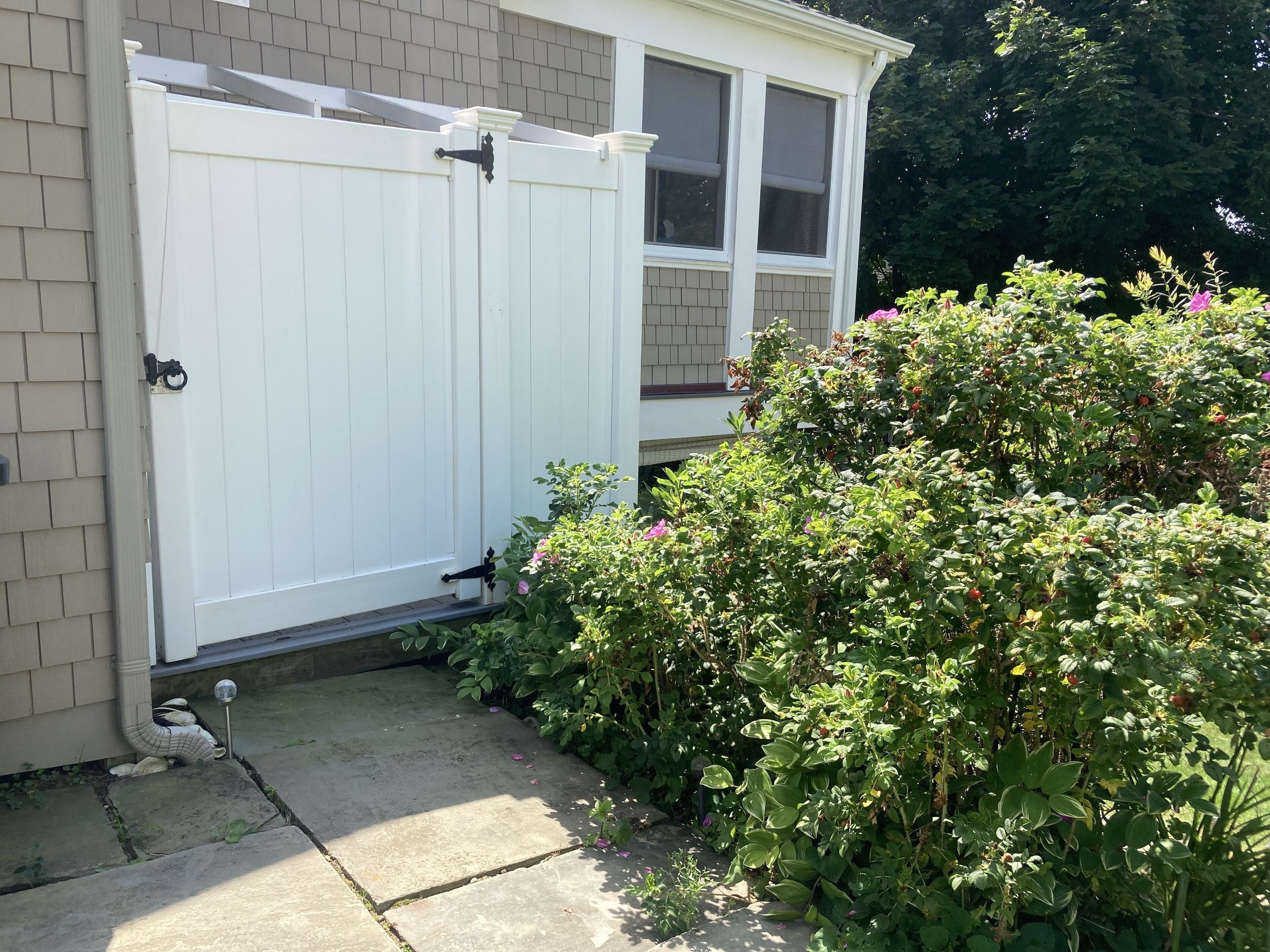 A white wooden gate with a metal latch, a brick house, and a lush green bush with pink flowers.