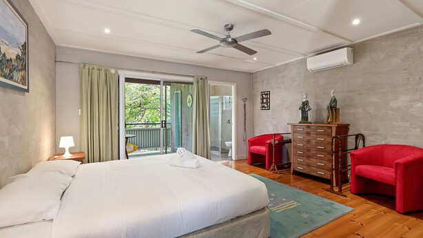 A bedroom with a bed, a ceiling fan, an air conditioning unit, a dresser, two red chairs, and a view of the outdoors through a sliding door.