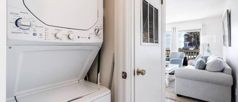 Un lavadero con lavadora y secadora de carga frontal, y una puerta que da a una sala de estar con sofá y lámpara.