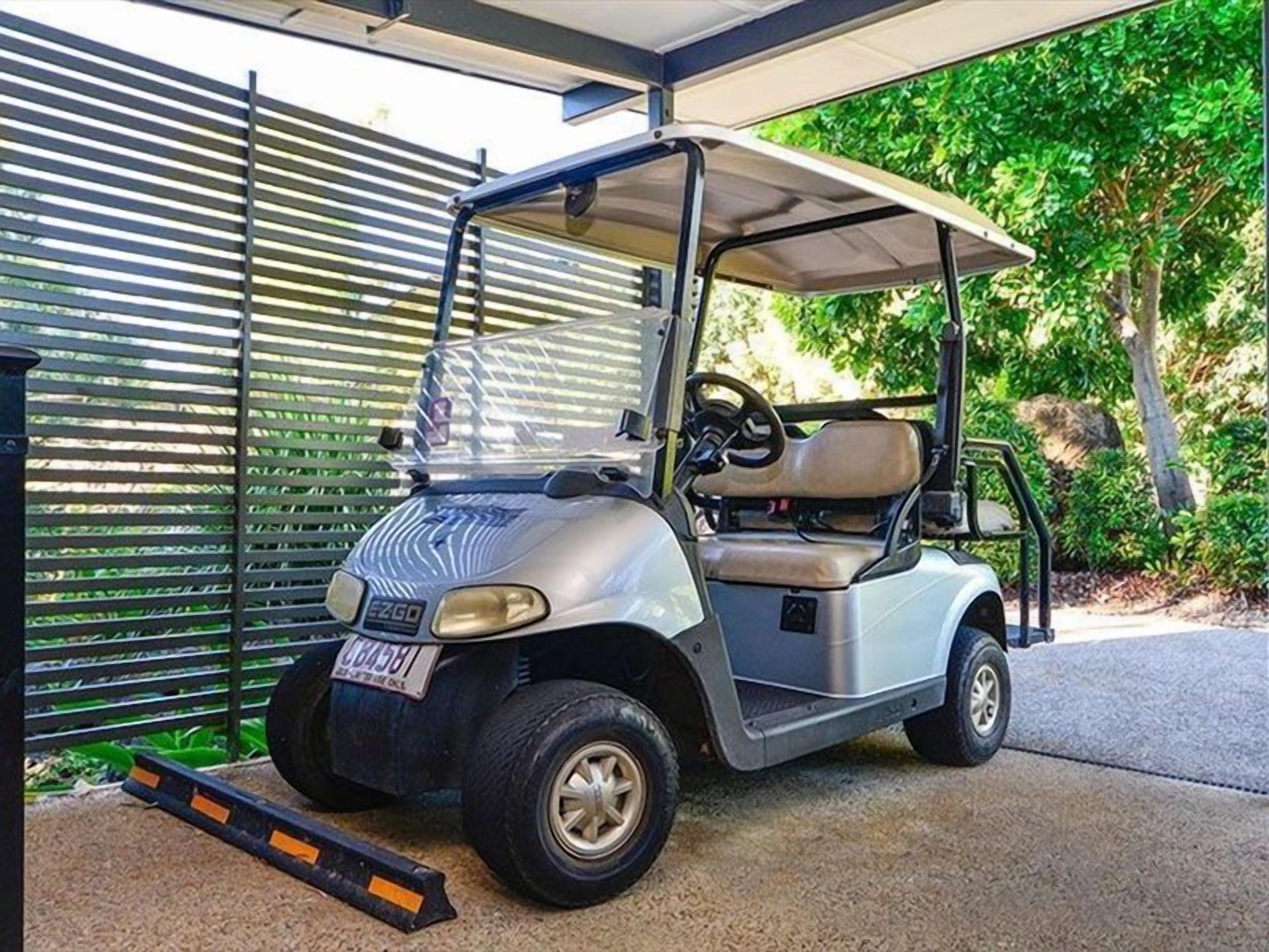 A golf cart parked in a covered area with a metal fence and greenery in the background.