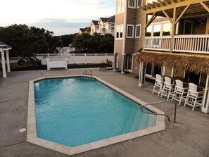 Una piscina rectangular con un deck blanco que la rodea, un área de bar techada con sillas blancas, y una cerca blanca al fondo.