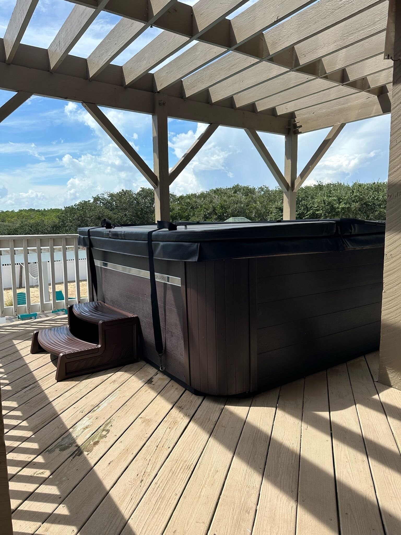 Een hottub onder een houten pergola op een houten terras, met uitzicht op bomen en een bewolkte lucht.