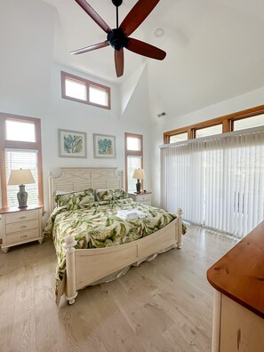 Un dormitorio con una cama grande, cabecera de madera, ventilador de techo y ventanas amplias con persianas.