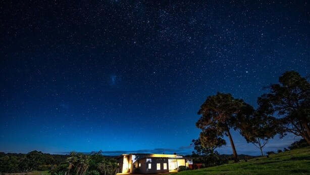 Uma casa sob um céu estrelado, cercada por árvores e uma colina gramada.