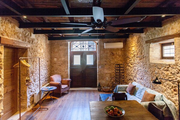 A rustic room with stone walls, wooden floors, a ceiling fan, a wooden table with a bowl of fruit, a sofa, a chair, and a radiator.