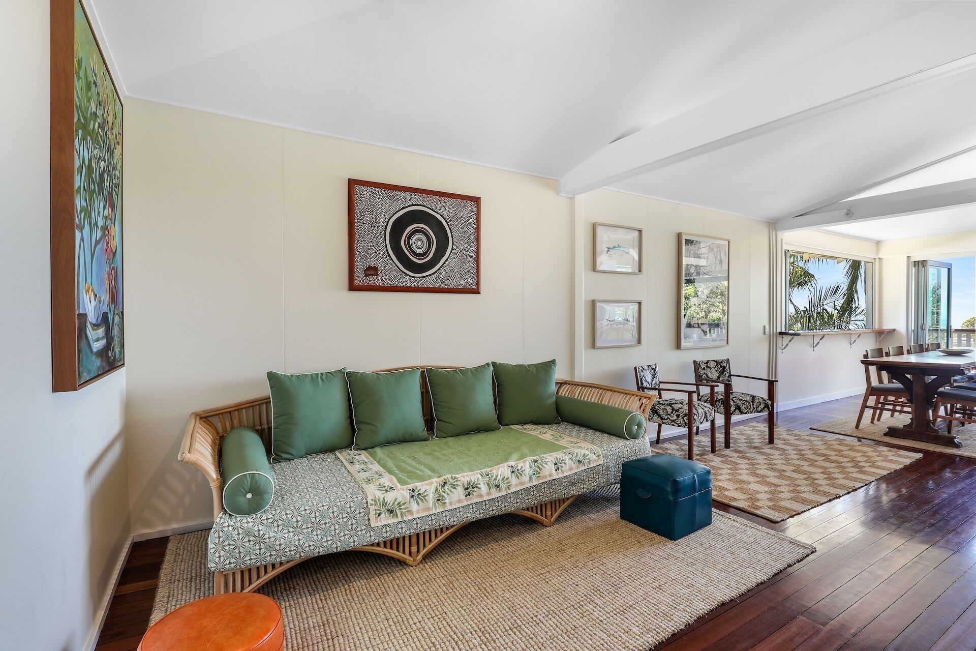A spacious living area with a sofa, a dining table, and chairs. There are framed artworks on the walls and a view of the outdoors through a large window.