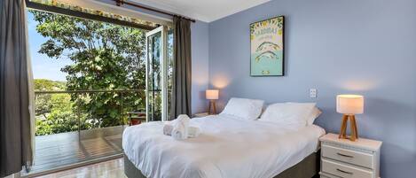 A bedroom with a large bed, two bedside tables with lamps, a ceiling fan, and a view of greenery through a large window.