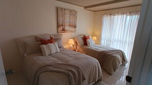 A hotel room with two beds, each with a pillow and a blanket, a painting on the wall, and a window with curtains.