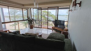 A spacious living room with a large glass window, a sofa, a coffee table, and a television. - Lowanna Unit 10, 24 Bulcock Beach Esplanade, Bulcock Beach (CALOUNDRA)