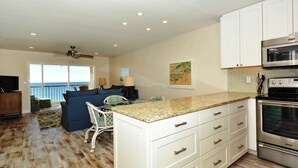 A spacious living area with a large window overlooking the ocean, a kitchen with white cabinets and stainless steel appliances, and a ceiling fan. - Siesta Key - Gulf-Front Retreat - 2 BR – Free Boat Docks - Upscale (Sarasota)