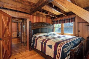 Une chambre à coucher rustique, avec un grand lit, un plafond en bois et une fenêtre ornée d’un rideau à carreaux.
