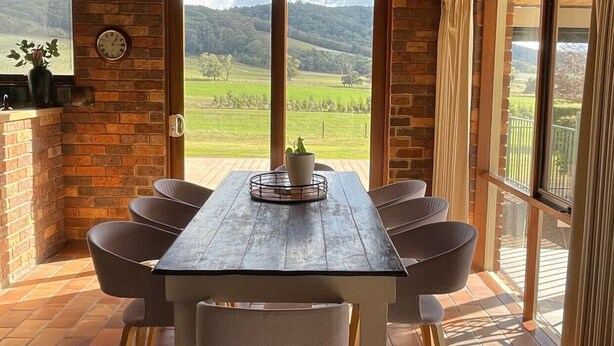 A rustic dining area with a wooden table and chairs, a potted plant, and a view of a green landscape through a large window. - Langbrook Estate Cottage (Yarra Junction)