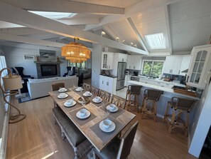 Une cuisine spacieuse avec des meubles blancs, un coin repas doté d’une table en bois pouvant accueillir six personnes, et un grand lustre.
