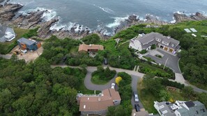 Vue aérienne d’une zone résidentielle côtière, avec des maisons, une côte rocheuse et un ciel dégagé.