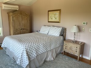 Un dormitorio con cama, mesita de noche, lámpara, armario, un cuadro en la pared y un termostato.