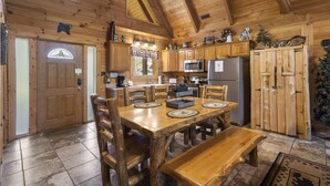 A wooden cabin interior with a dining area, kitchen, and a large wooden door.