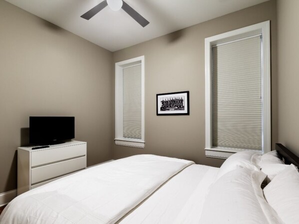 A bedroom with a bed, a dresser, a television, and two windows with blinds.